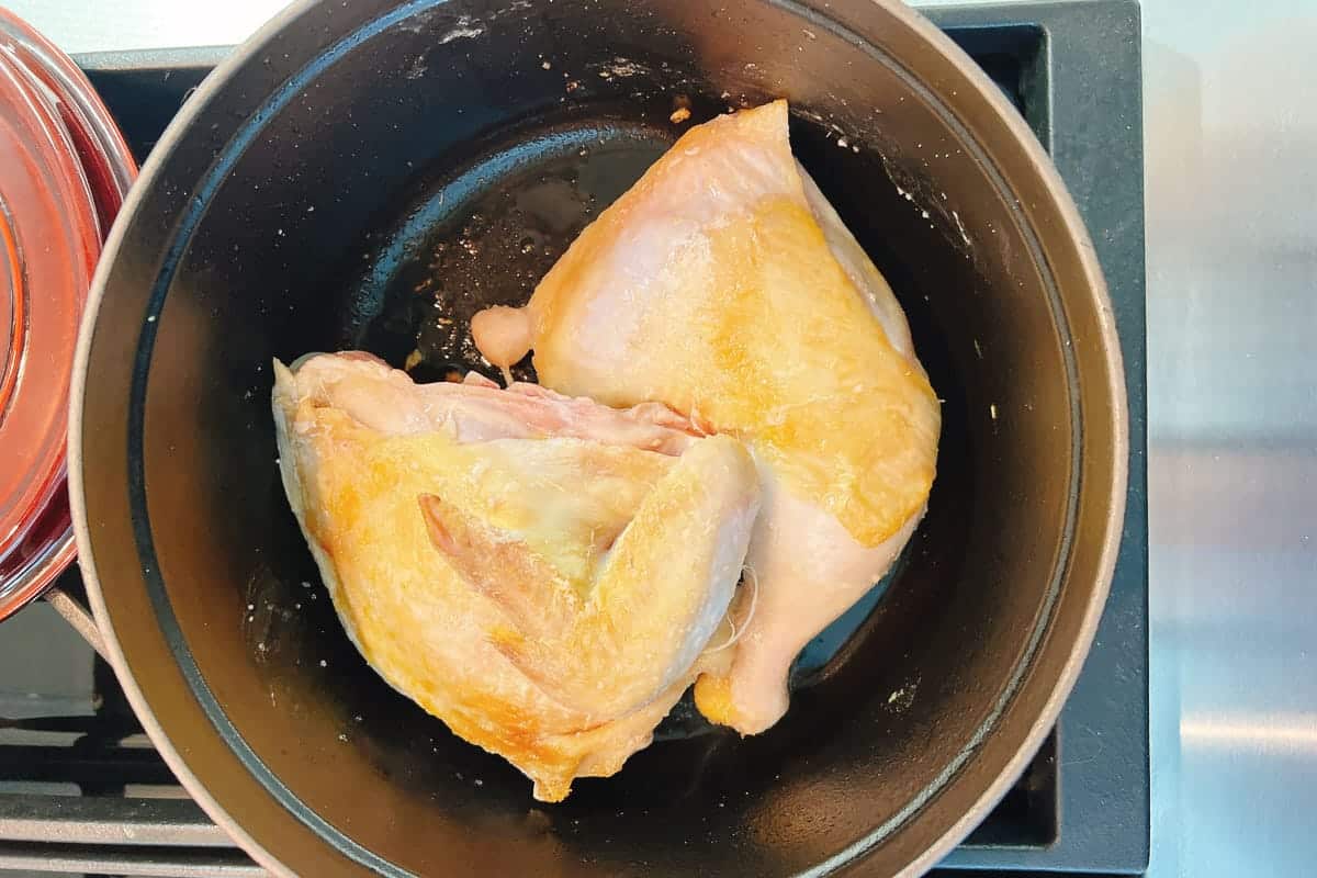Two bone-in, skin-on chicken breasts and leg quarters are being browned in a black pot on a stovetop—perfectly golden and crispy, ready to be simmered into a comforting chicken ginger soup.
