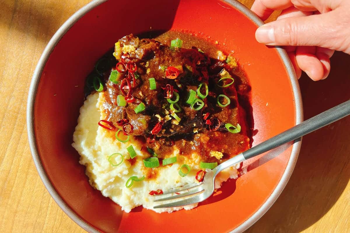 A hand holds a red bowl filled with creamy mashed potatoes topped with braised short ribs, savory sauce, sliced green onions, and red chili peppers, with a fork resting inside.