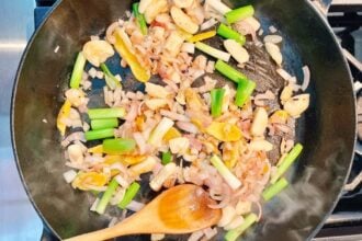 Chopped onions, green onions, garlic, and yellow bell peppers sizzle in a black skillet as a wooden spoon stirs. Steam rises from the pan—perfect aromatics for starting flavorful braised short ribs.