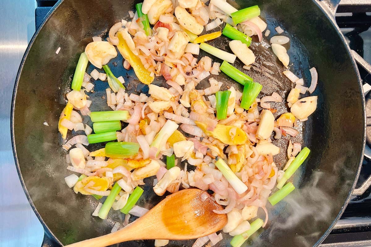 Chopped onions, green onions, garlic, and yellow bell peppers sizzle in a black skillet as a wooden spoon stirs. Steam rises from the pan—perfect aromatics for starting flavorful braised short ribs.