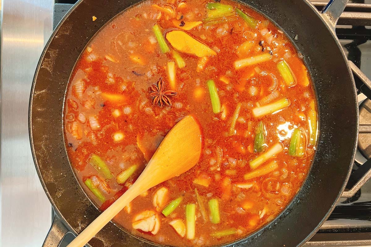 A pan of simmering broth with braised short ribs, chopped green onions, garlic cloves, ginger slices, star anise, and a wooden spoon on a stovetop. The reddish-brown broth is rich with aromatic spices, vegetables, and tender meat.
