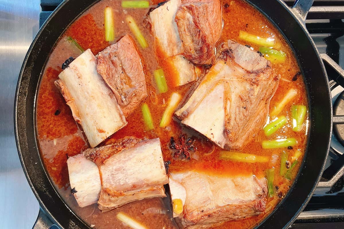 Braised short ribs simmer in a reddish-brown miso ginger broth with green onion pieces in a black pot on the stovetop. The rich, flavorful broth surrounds the tender ribs, which are partially submerged.