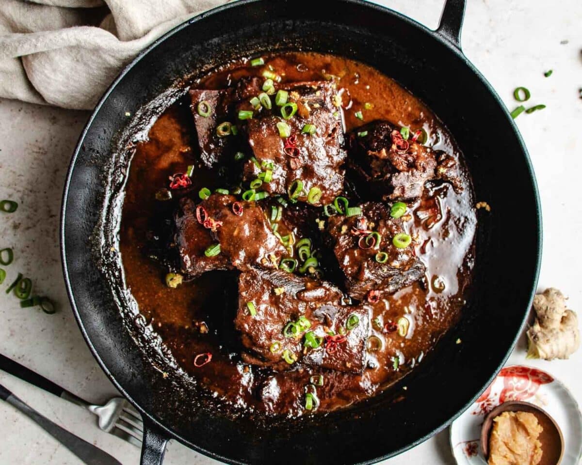 Braised short ribs in a rich, dark miso ginger sauce are served in a black cast iron pan, garnished with chopped green onions and red chili slices. A fork, knife, ginger, and miso paste complete this hearty braised short ribs presentation.
