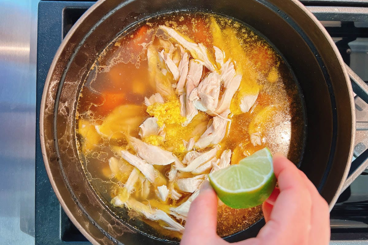 A hand squeezes a lime over a pot of chicken ginger soup, filled with shredded chicken, broth, and grated garlic simmering on the stovetop.