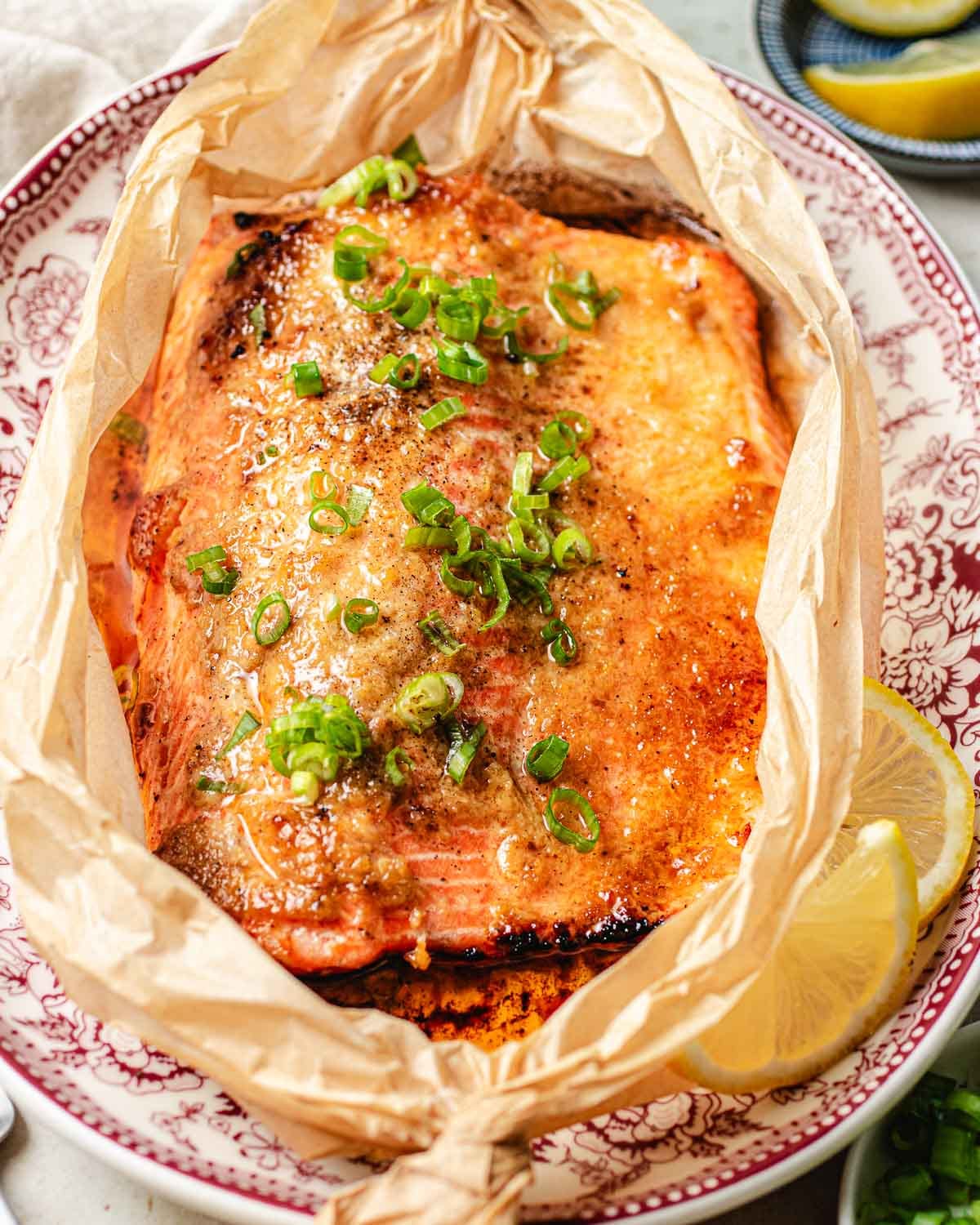 A baked rainbow trout fillet with chopped green onions sits on a decorative red and white plate, wrapped in parchment paper, with a lemon wedge on the side.