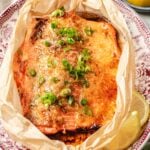 A baked trout topped with miso butter and chopped green onions sits on parchment paper on a decorative plate, with a lemon wedge on the side.