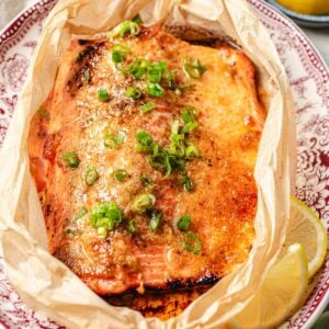 A baked trout topped with miso butter and chopped green onions sits on parchment paper on a decorative plate, with a lemon wedge on the side.