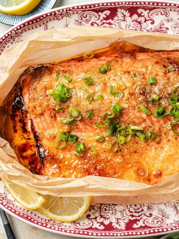 A baked trout topped with chopped green onions and flavored with miso and butter sits on parchment paper, garnished with lemon slices, on a decorative red and white plate.