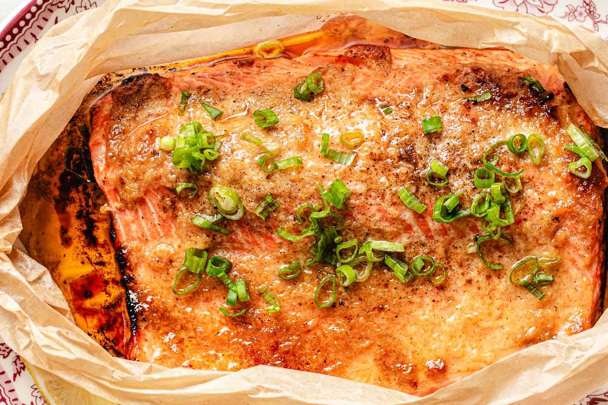 A baked trout fillet topped with chopped green onions and spices, served on parchment paper with browned edges and a light miso butter glaze on top.