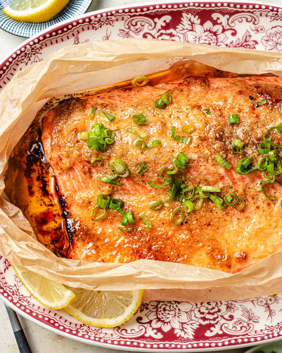 A baked trout topped with chopped green onions and flavored with miso and butter sits on parchment paper, garnished with lemon slices, on a decorative red and white plate.