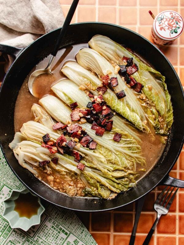A cast iron pan filled with braised napa cabbage topped with crispy pieces of bacon, all nestled in a savory brown sauce. A silver ladle rests in the pan, which sits on an orange tiled surface beside a patterned cloth and condiment jar.