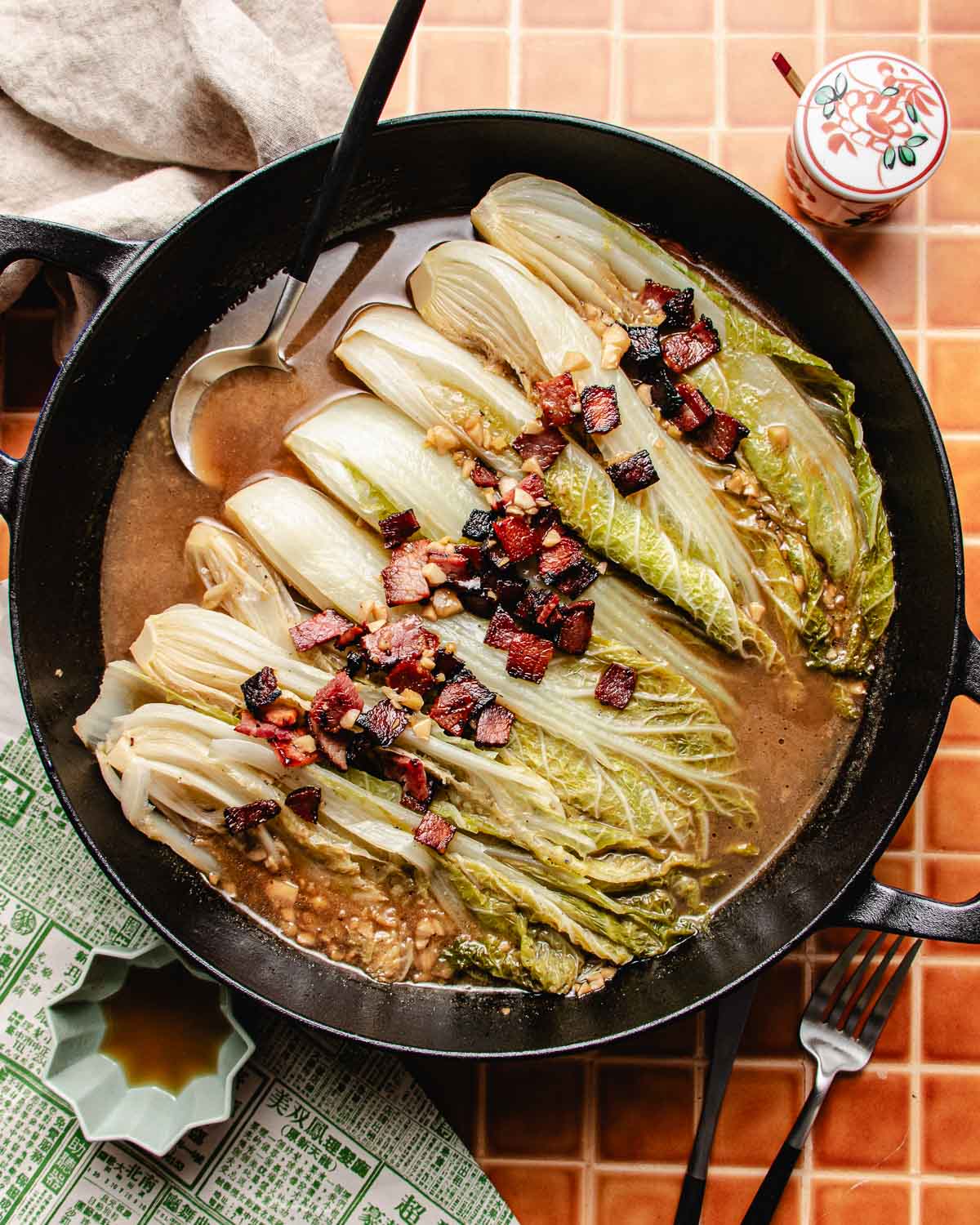 A cast iron pan filled with braised napa cabbage topped with crispy pieces of bacon, all nestled in a savory brown sauce. A silver ladle rests in the pan, which sits on an orange tiled surface beside a patterned cloth and condiment jar.
