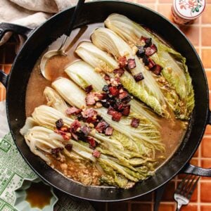 A cast iron skillet filled with braised napa cabbage halves in savory broth, topped with crispy bacon pieces, sits on a tiled surface with a serving spoon resting inside.