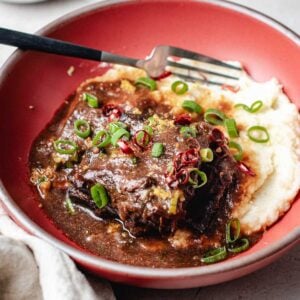 A red bowl with mashed potatoes topped with braised short ribs in a rich miso ginger sauce, garnished with sliced green onions and red chili, with a fork resting on the side.