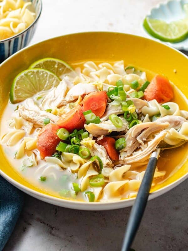 A bowl of chicken ginger soup with carrots, shredded chicken, egg noodles, green onions, and lime slices, served with a spoon. A bowl of plain noodles and lime wedges are in the background.