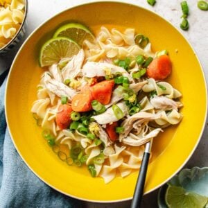 A yellow bowl of chicken ginger soup with egg noodles, shredded chicken, carrot slices, chopped green onions, lime wedges, and a black-handled spoon, set on a light surface with a blue napkin.