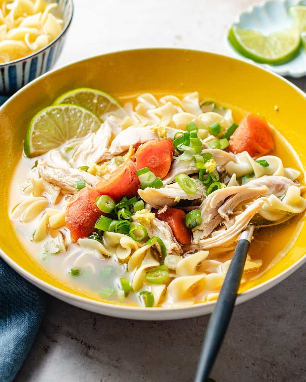 A bowl of chicken ginger soup with carrots, shredded chicken, egg noodles, green onions, and lime slices, served with a spoon. A bowl of plain noodles and lime wedges are in the background.