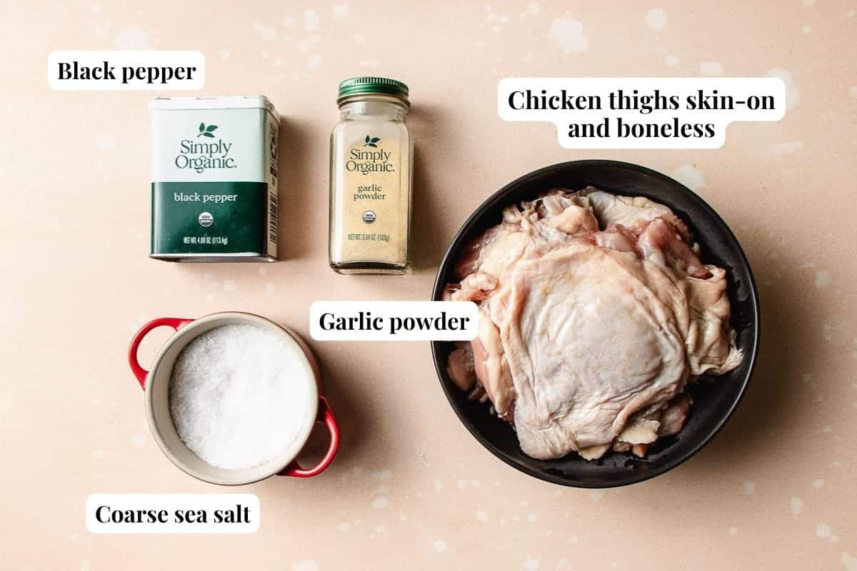 A bowl of raw, skin-on, boneless chicken thighs sits next to containers of black pepper, garlic powder, and coarse sea salt—perfect for preparing flavorful honey garlic chicken thighs.