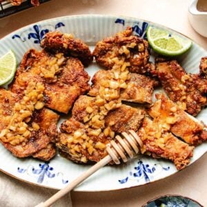 Crispy chicken thighs, reminiscent of honey garlic chicken thighs, are topped with diced garlic on a decorative plate, garnished with lime wedges. A honey dipper rests beside, hinting at the dish’s signature honey garlic flavor.