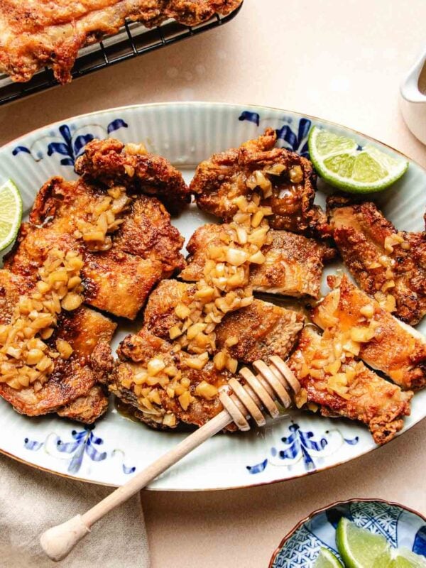 A platter of crispy honey garlic chicken thighs, topped with diced garlic and garnished with lime wedges. A honey dipper rests on the plate, suggesting a sweet drizzle over the chicken on a decorative blue and white plate.
