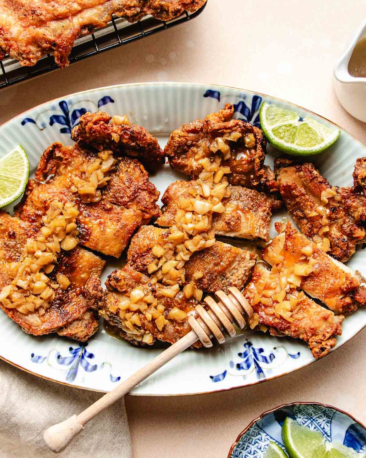 A platter of crispy honey garlic chicken thighs, topped with diced garlic and garnished with lime wedges. A honey dipper rests on the plate, suggesting a sweet drizzle over the chicken on a decorative blue and white plate.