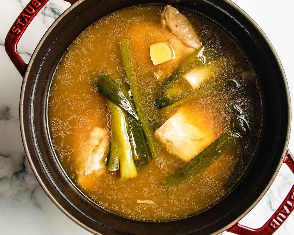 A pot of chicken ginger soup with chicken pieces, green onions, and ginger slices in a light brown broth, seen from above. The black pot with red handles rests on a white marble surface.