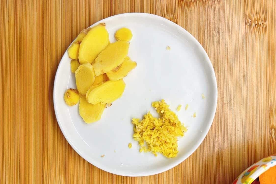 A white plate on a wooden surface holds sliced fresh ginger on the left and a small pile of grated ginger on the right, perfect for adding to your favorite chicken ginger soup recipe.