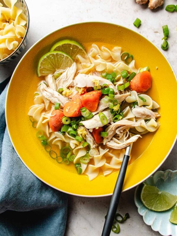 A yellow bowl filled with chicken noodle soup with ginger, sliced carrots, green onions, egg noodles, and lime wedges, with a spoon resting in the bowl and a blue napkin beside it. Extra noodles and lime slices are nearby.