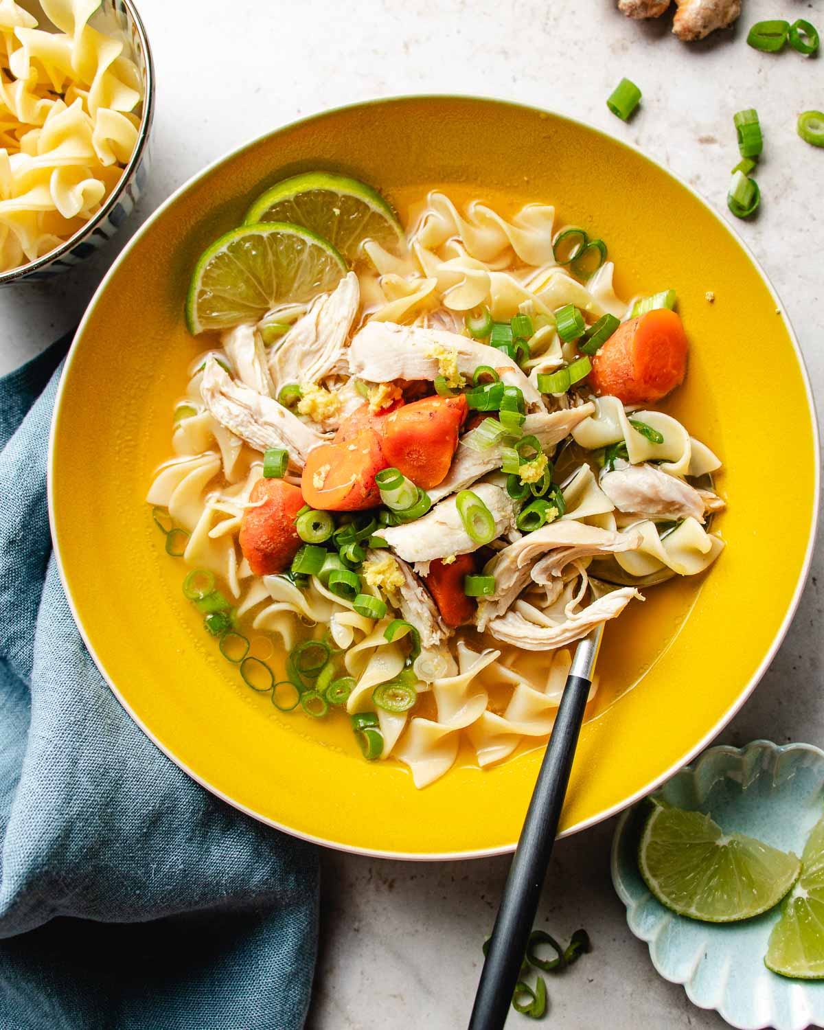 A yellow bowl filled with chicken noodle soup with ginger, sliced carrots, green onions, egg noodles, and lime wedges, with a spoon resting in the bowl and a blue napkin beside it. Extra noodles and lime slices are nearby.