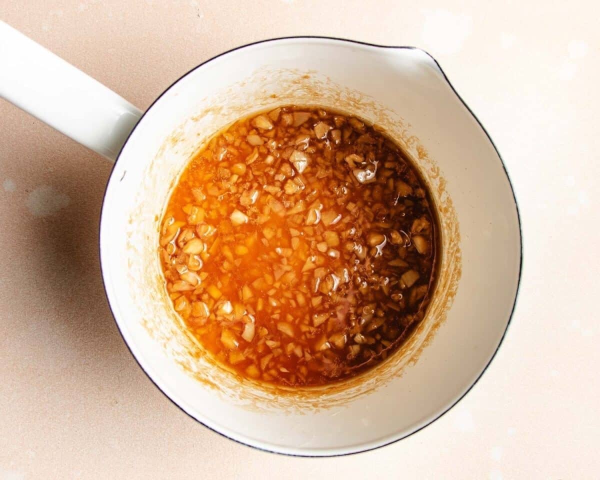 A white saucepan contains a golden-brown mixture of honey garlic butter sauce with finely chopped garlic in oil, the savory base for delicious honey garlic chicken thighs, sitting on a light-colored surface.
