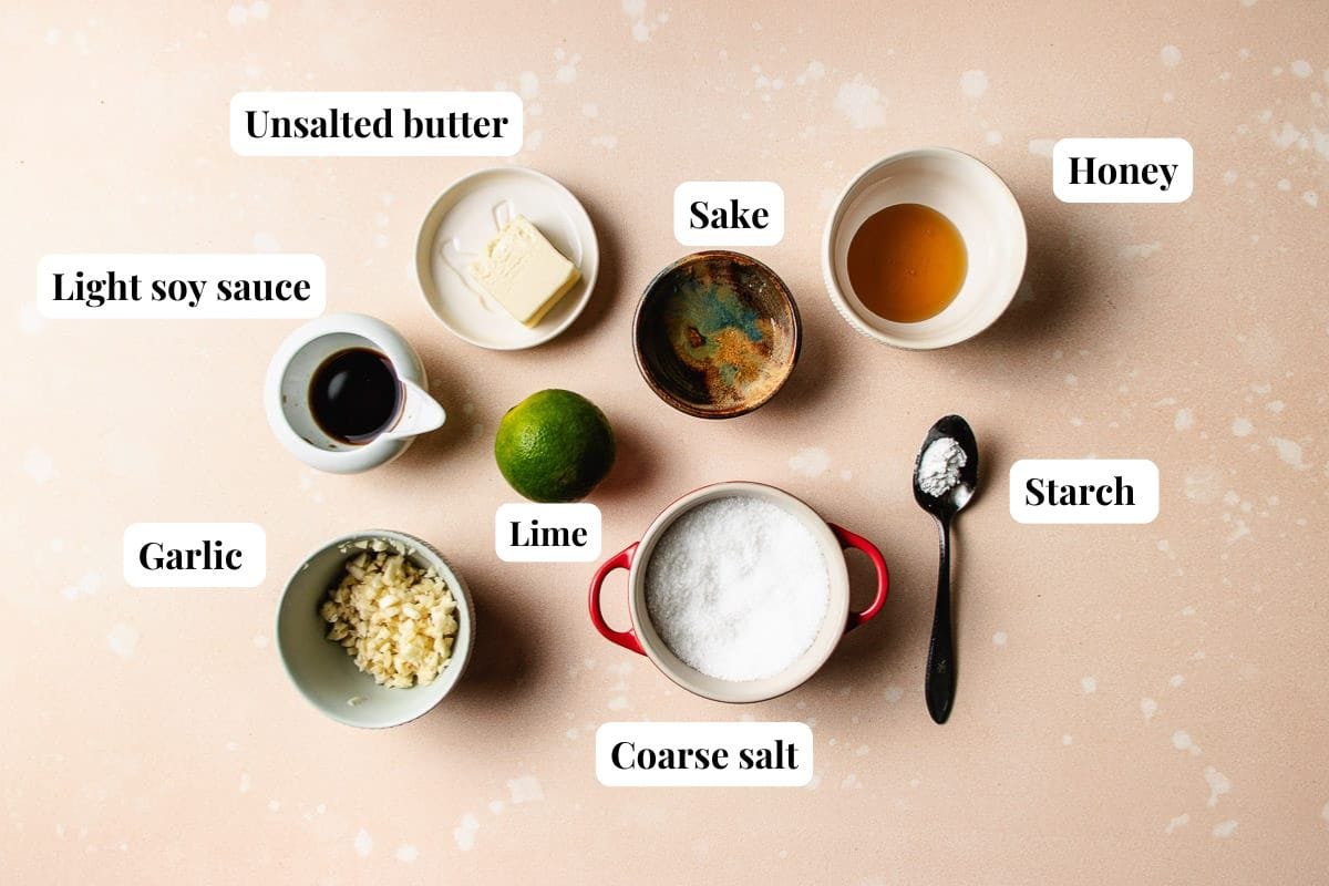 A flat lay of ingredients for honey garlic chicken thighs—unsalted butter, honey, sake, light soy sauce, garlic, lime, coarse salt, and starch—arranged in bowls and containers on a light pink speckled surface.