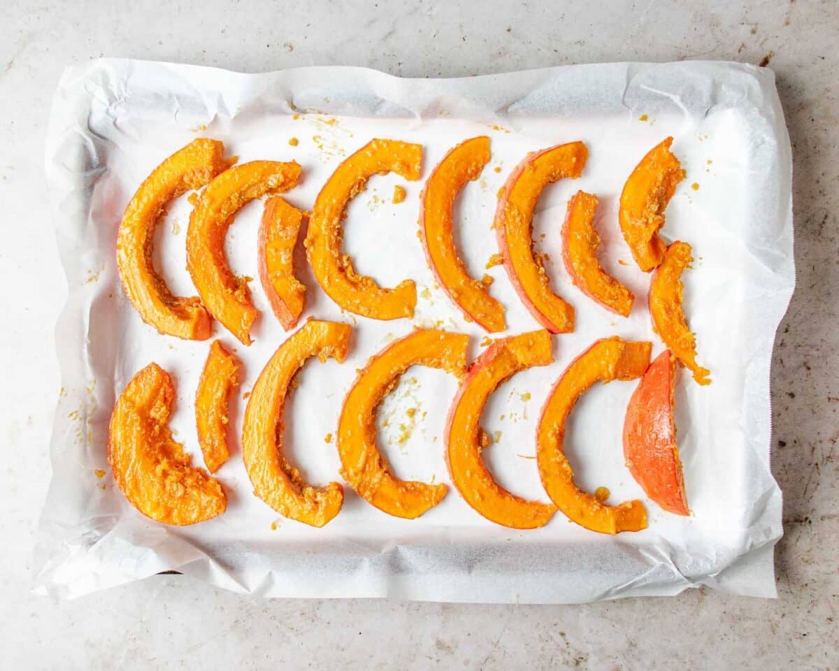 Slices of raw roasted red kuri squash coated with seasoning are arranged in rows on a parchment-lined baking tray, ready for roasting. The tray sits on a light-colored countertop.