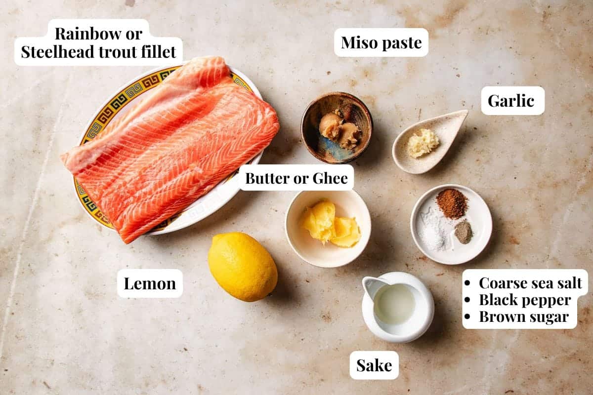 A flat lay of ingredients for baked trout, featuring a fillet of rainbow or steelhead trout, miso paste, garlic, lemon, butter or ghee, sake, coarse sea salt, black pepper, and brown sugar, all labeled.