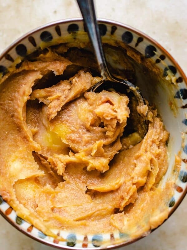 A bowl filled with creamy, light brown miso butter, partially mixed with a spoon, sits on a light-colored surface.