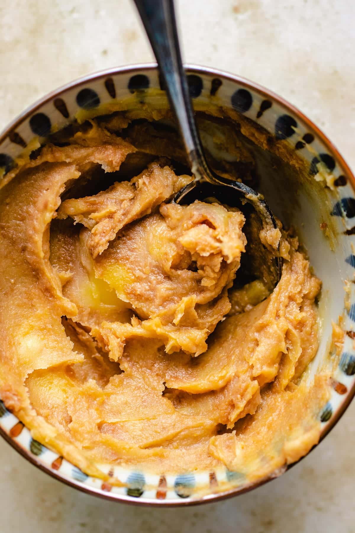 A bowl filled with creamy, light brown miso butter, partially mixed with a spoon, sits on a light-colored surface.