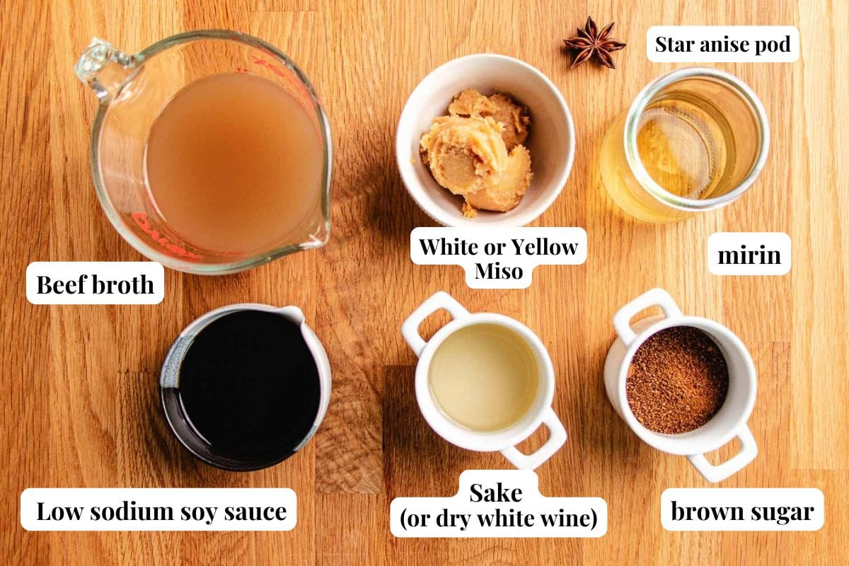 Seven labeled ingredients for braised short ribs sauce are arranged on a wooden surface: beef broth, low sodium soy sauce, white or yellow miso, sake (or dry white wine), brown sugar, mirin, and a star anise pod.