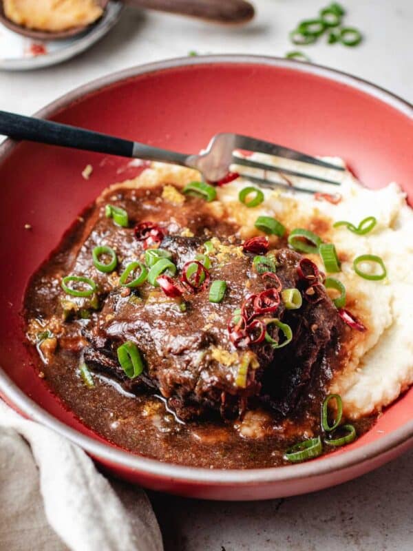 A red bowl with braised short ribs in rich brown miso ginger sauce, topped with sliced green onions and red chili, served alongside creamy mashed potatoes. A fork rests in the bowl, ready to enjoy the tender braised short ribs.
