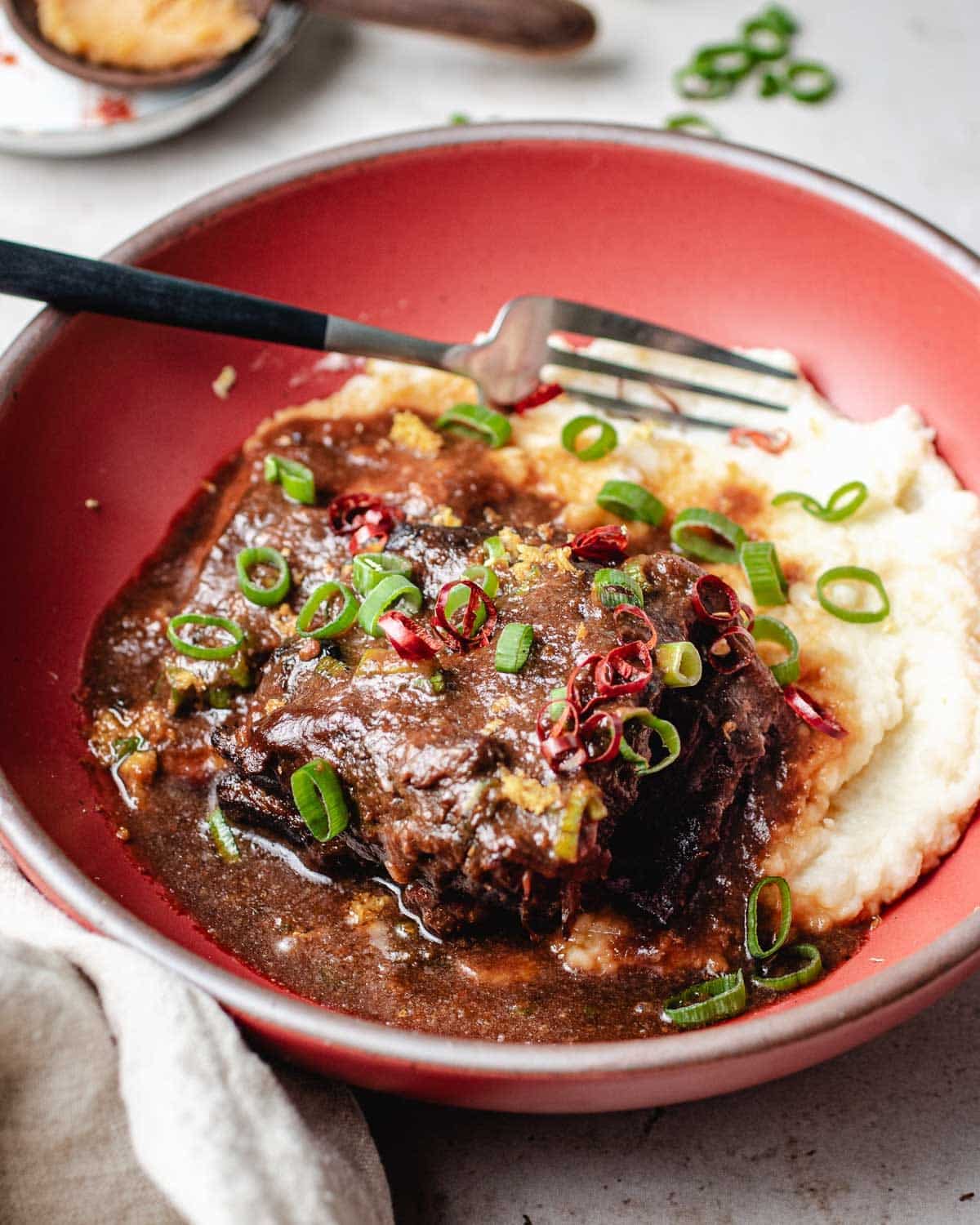 A red bowl with braised short ribs in rich brown miso ginger sauce, topped with sliced green onions and red chili, served alongside creamy mashed potatoes. A fork rests in the bowl, ready to enjoy the tender braised short ribs.