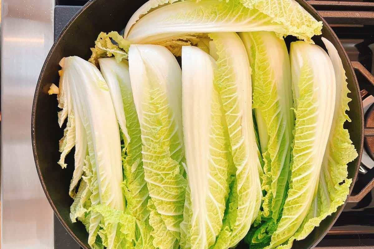 Several long, fresh napa cabbage leaves are arranged side by side in a black pan on a stovetop, ready to be cooked for a delicious braised napa cabbage dish.