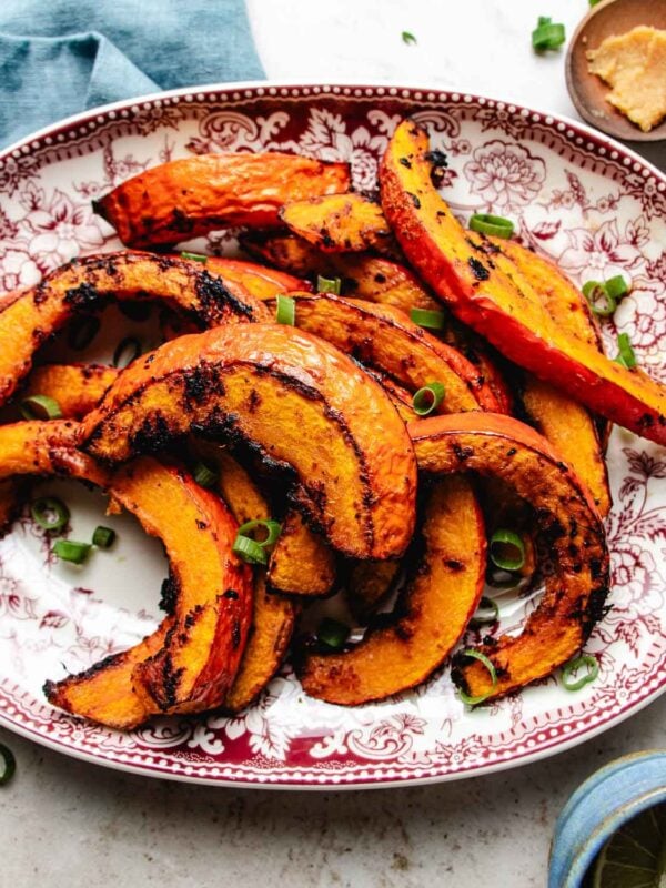 Slices of roasted red kuri squash with browned edges, garnished with chopped green onions, are arranged on a red and white patterned plate.