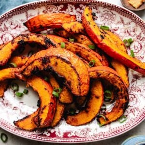 Slices of roasted red kuri squash with char marks are arranged on a decorative red and white platter, garnished with chopped green onions.