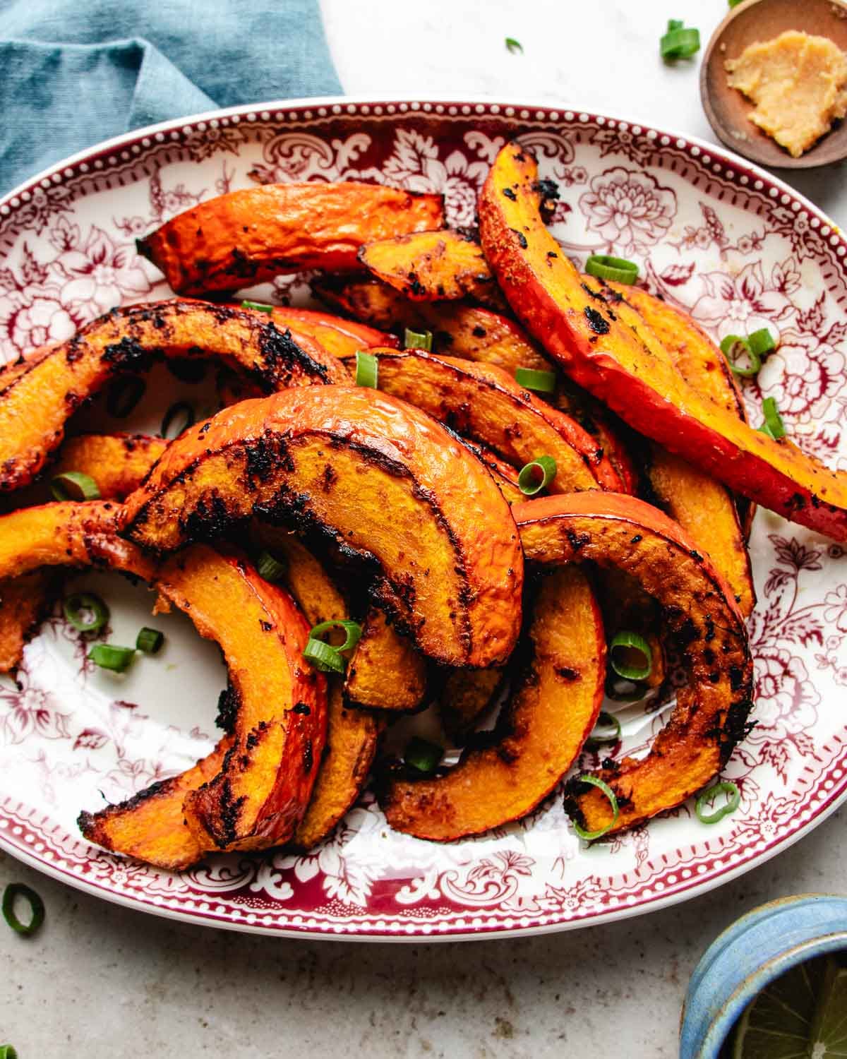 Slices of roasted red kuri squash with browned edges, garnished with chopped green onions, are arranged on a red and white patterned plate.