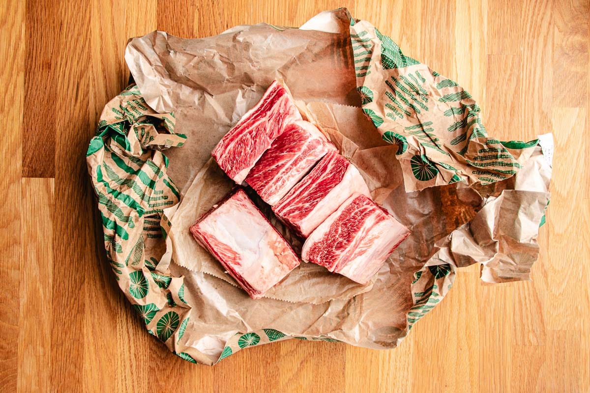 Four raw beef short ribs with visible marbling are placed on brown butcher paper with green patterns, set on a wooden surface—perfect for preparing rich, flavorful braised short ribs.