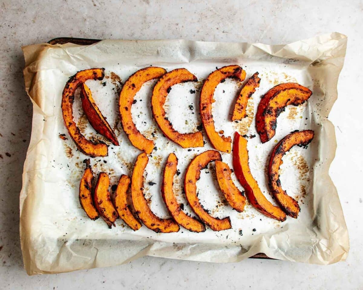Sliced roasted red kuri squash arranged on a parchment-lined baking sheet. The slices are golden brown with caramelized edges, sprinkled with herbs and seasoning.