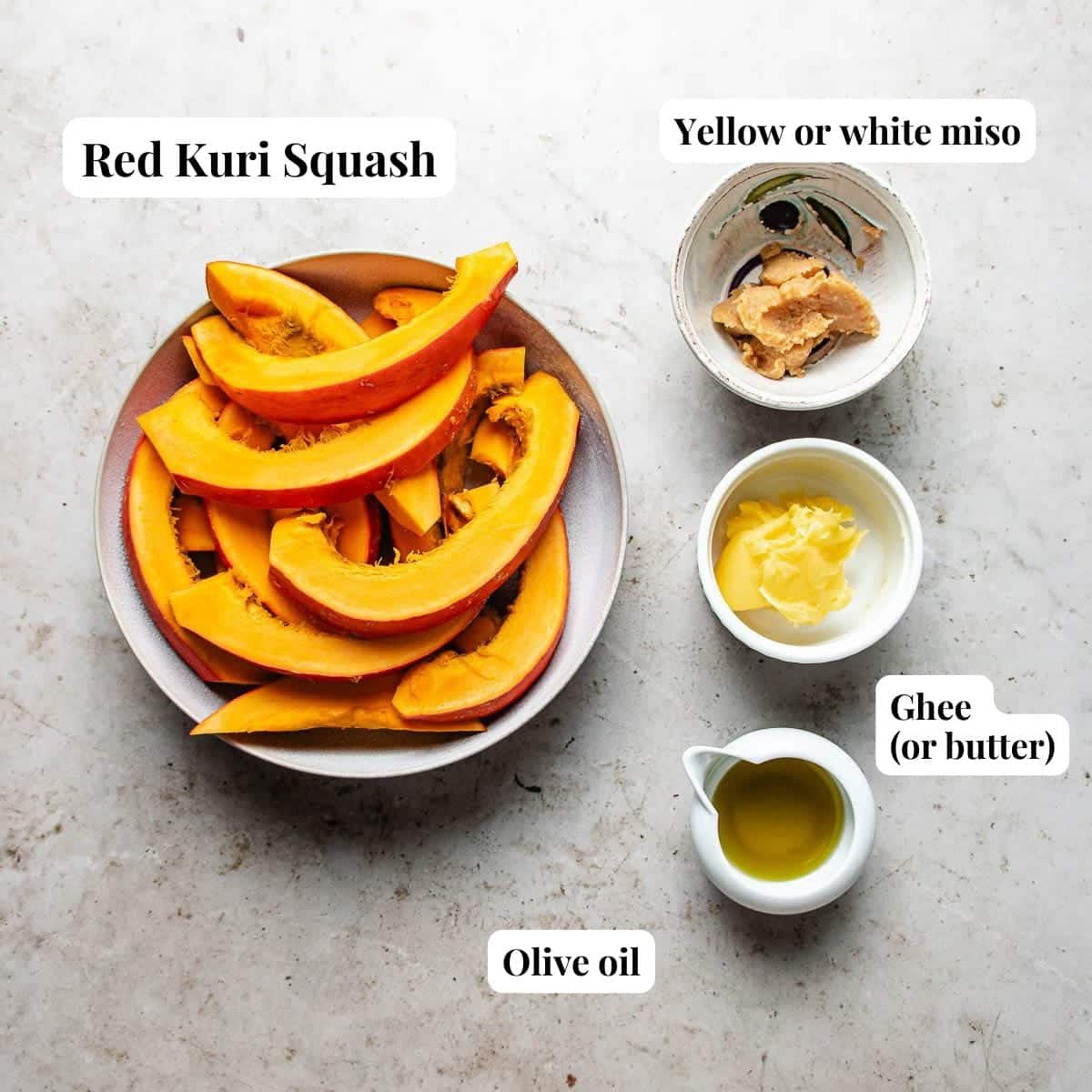 Sliced roasted red kuri squash in a bowl, with small bowls of yellow or white miso, ghee (or butter), and olive oil arranged beside it on a light surface. Each ingredient is labeled.