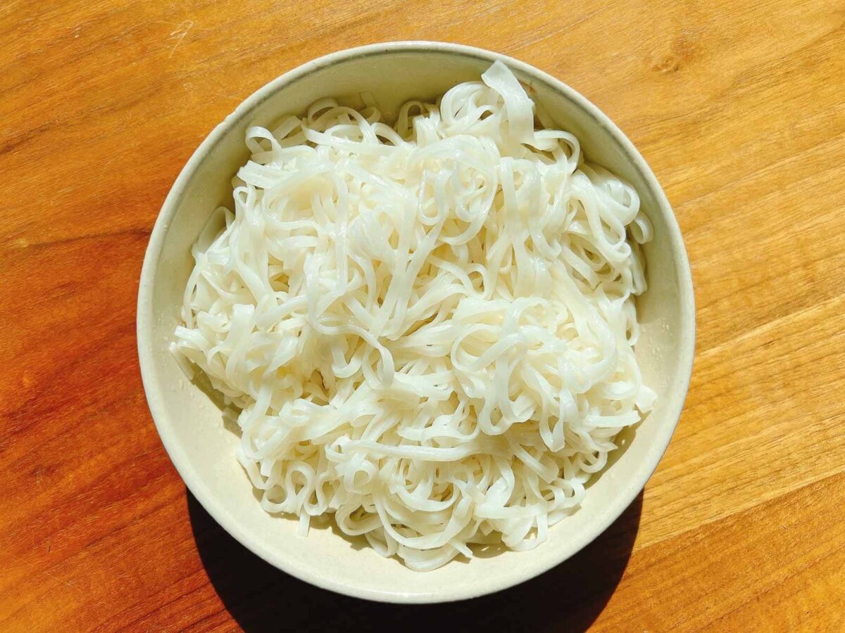 A bowl filled with plain cooked white rice pho noodles sits on a wooden surface, brightly lit by natural sunlight, ready to be paired with flavorful turkey pho bone broth.