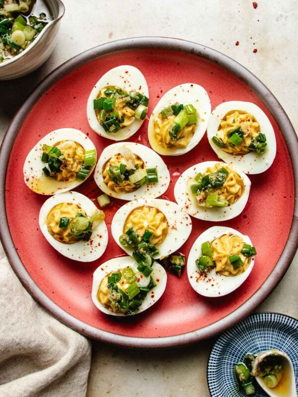 A red plate holds nine Asian deviled eggs topped with chopped green onions and herbs, sprinkled with red spices. A bowl of more chopped greens and a small blue dish are nearby on a light-colored surface.