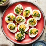 A red plate with nine Asian deviled eggs, topped with chopped green onions, scallion oil, and sprinkled with red seasoning, sits on a neutral-colored surface.