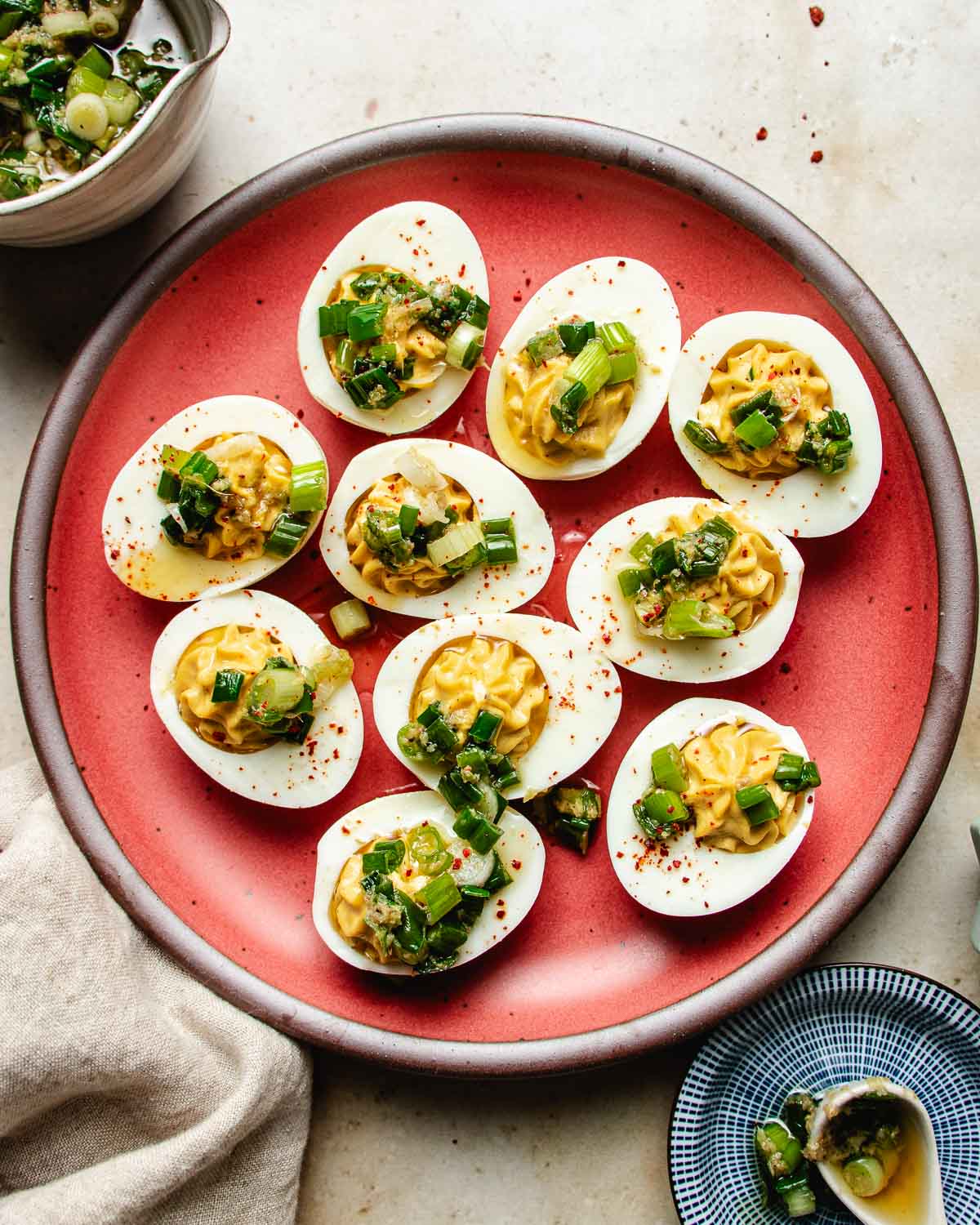 A red plate holds nine Asian deviled eggs topped with chopped green onions and herbs, sprinkled with red spices. A bowl of more chopped greens and a small blue dish are nearby on a light-colored surface.