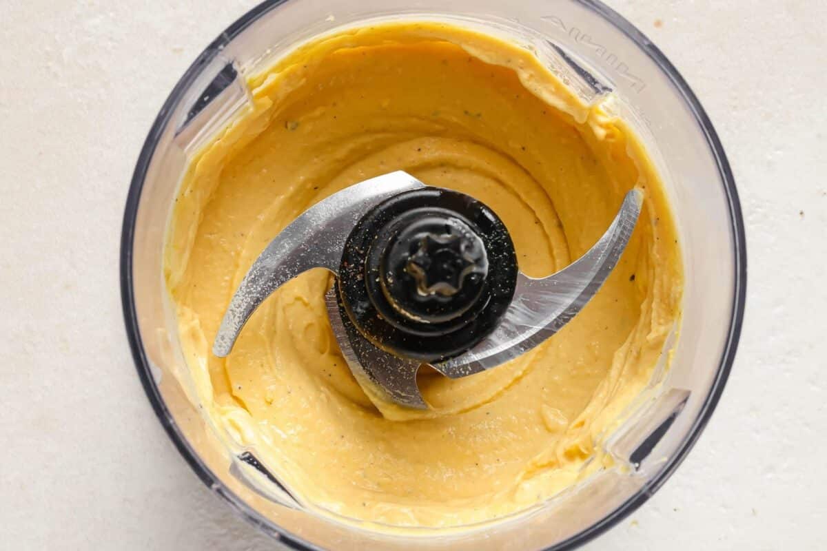 A food processor viewed from above, containing a smooth, creamy egg yolk blend mixture for Asian deviled eggs, with two sharp metal blades in the center.